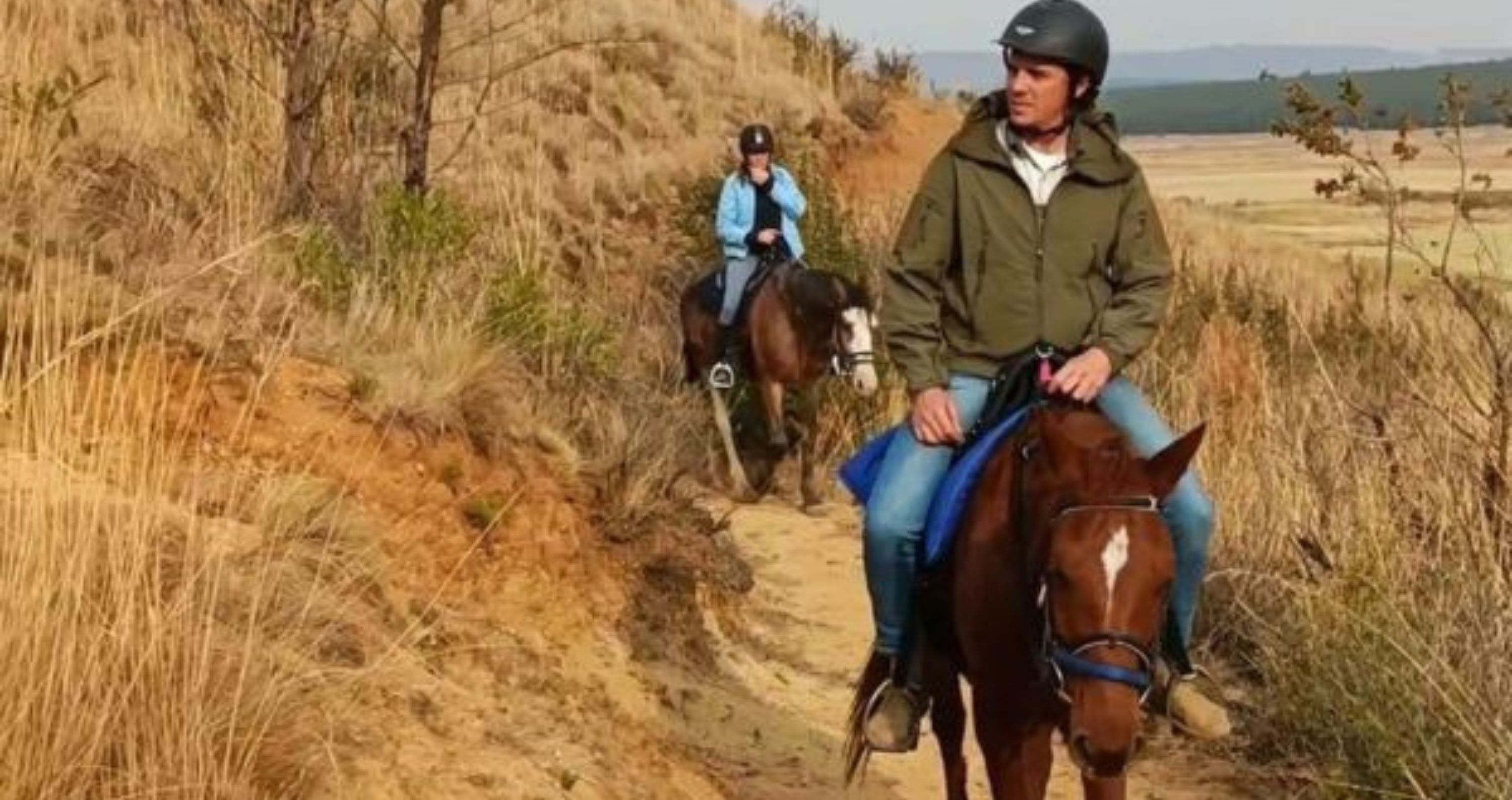 Dos personas montando a caballo por un sendero de tierra rodeado de hierba seca y escasos árboles.
