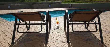 A poolside area with two chairs facing a pool, a glass of drink, and a view of a hilly landscape.