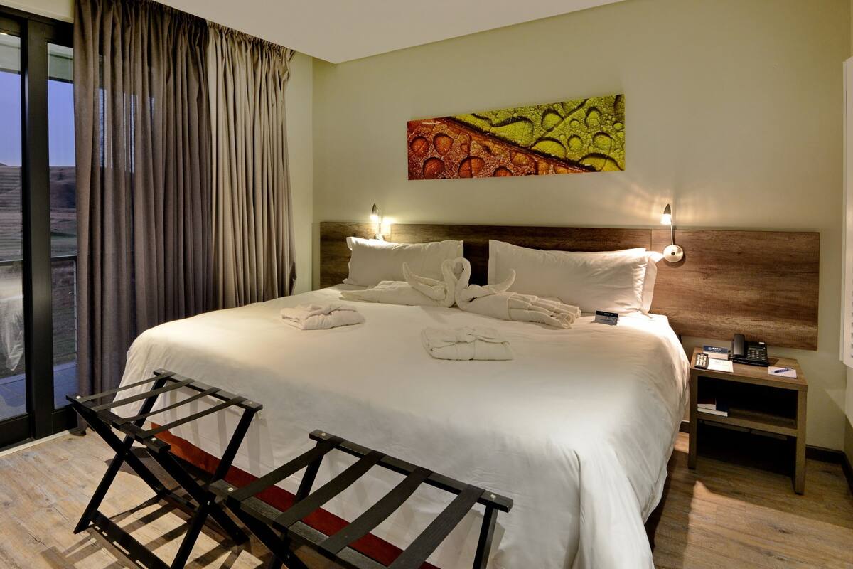 Ein Hotelzimmer mit einem großen Bett, Blick durch eine Schiebetür, einem Bild an der Wand und einem Nachttisch mit Lampe.