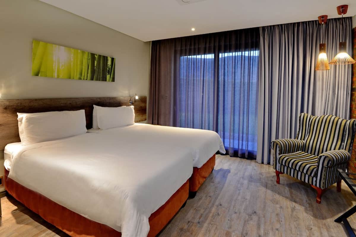 Ein Hotelzimmer mit einem großen Bett, einem hölzernen Kopfteil, einem gestreiften Sessel und Blick ins Freie durch ein Fenster mit Vorhängen.