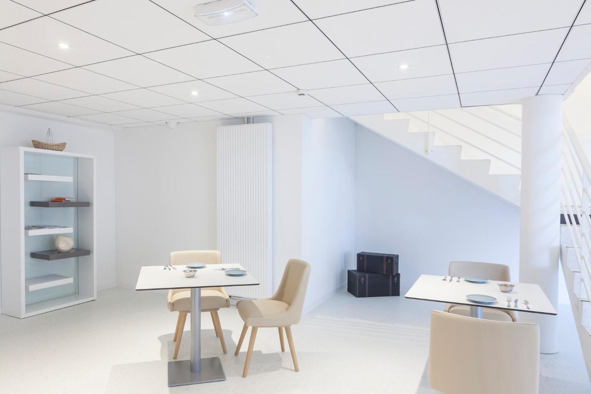 A modern, well-lit office space with two tables and chairs, a white bookshelf, and a staircase.