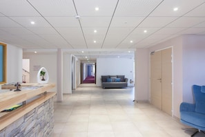 A modern hotel lobby with a reception desk, a seating area with a sofa, and a corridor leading to other rooms. - Hôtel Spa Les Rives Sauvages (Malbuisson)
