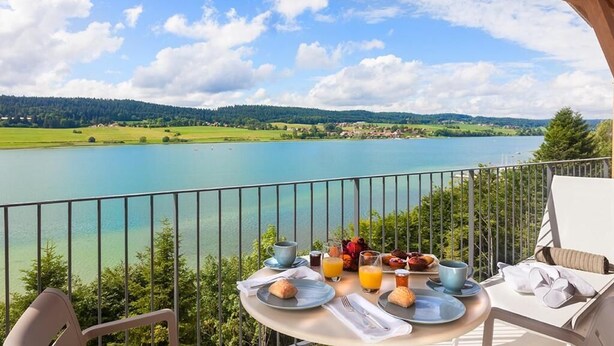 Un balcon avec une table dressée pour le petit-déjeuner, donnant sur un lac et des collines.