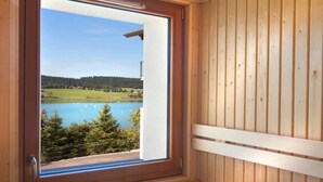 Un sauna en bois doté d’une fenêtre donnant sur un lac et le paysage environnant.