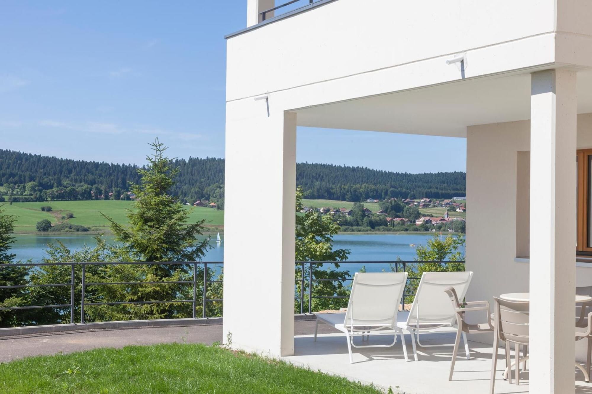 Een modern terras buiten met witte stoelen en een tafel, met uitzicht op een meer en beboste heuvels.