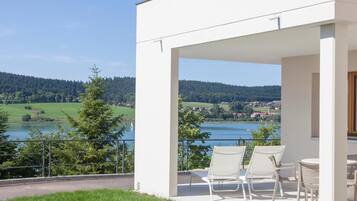 Een modern terras buiten met witte stoelen en een tafel, met uitzicht op een meer en beboste heuvels.