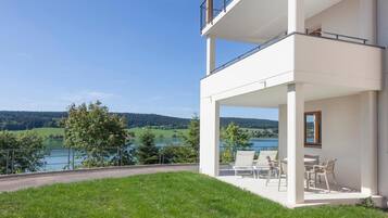 A modern two-story building with a balcony overlooking a lake and forested hills.