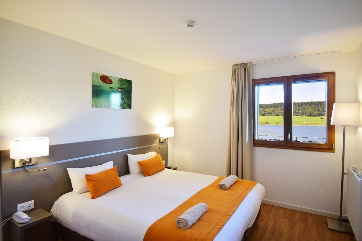 Ein Hotelzimmer mit einem großen Bett, zwei Nachttischlampen, einem Gemälde an der Wand, einem Fenster mit Seeblick und einer Stehlampe.