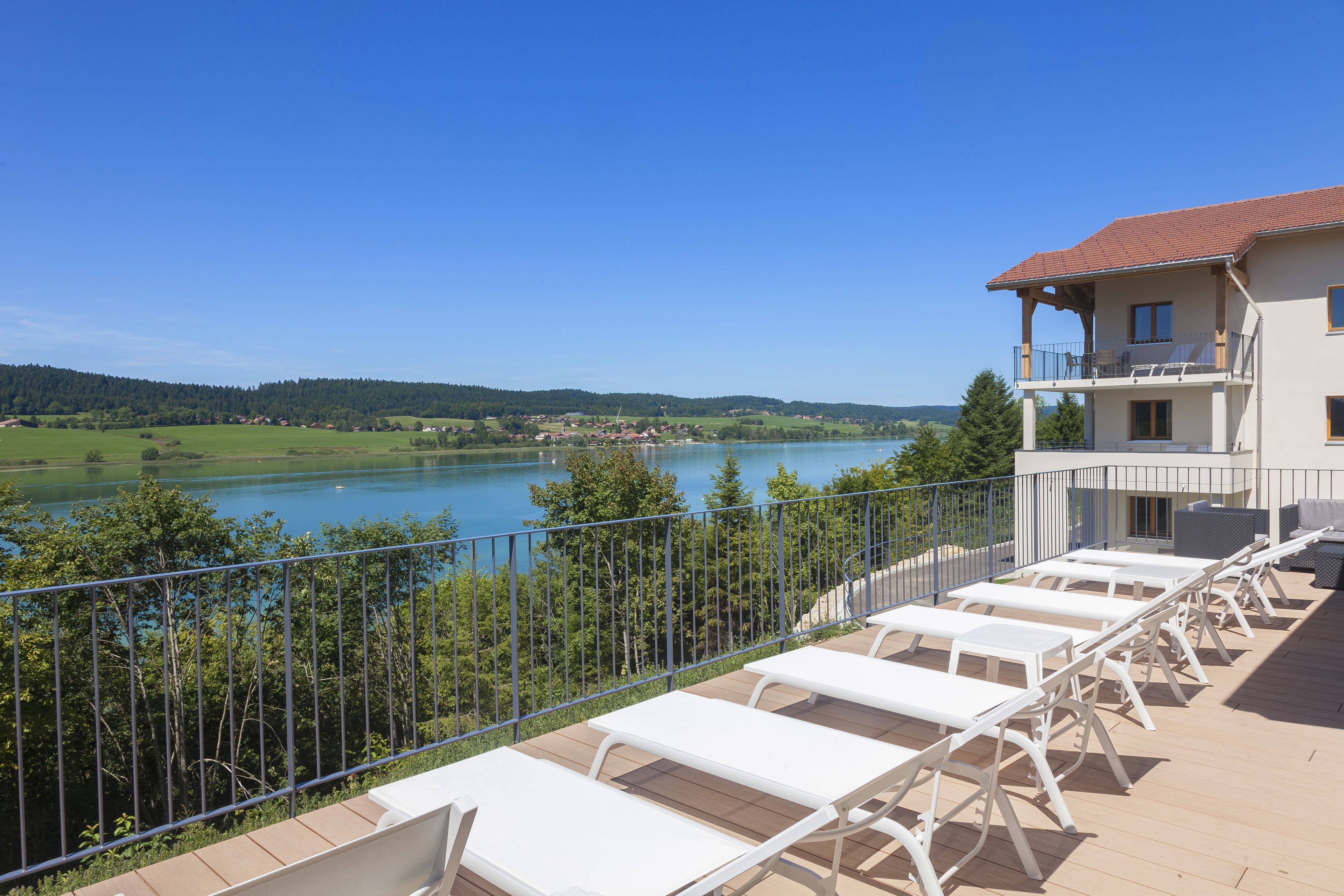 A terrace with white sun loungers overlooking a lake, a building with a balcony, and a clear blue sky.