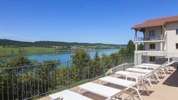 A terrace with white sun loungers overlooking a lake, a building with a balcony, and a clear blue sky.