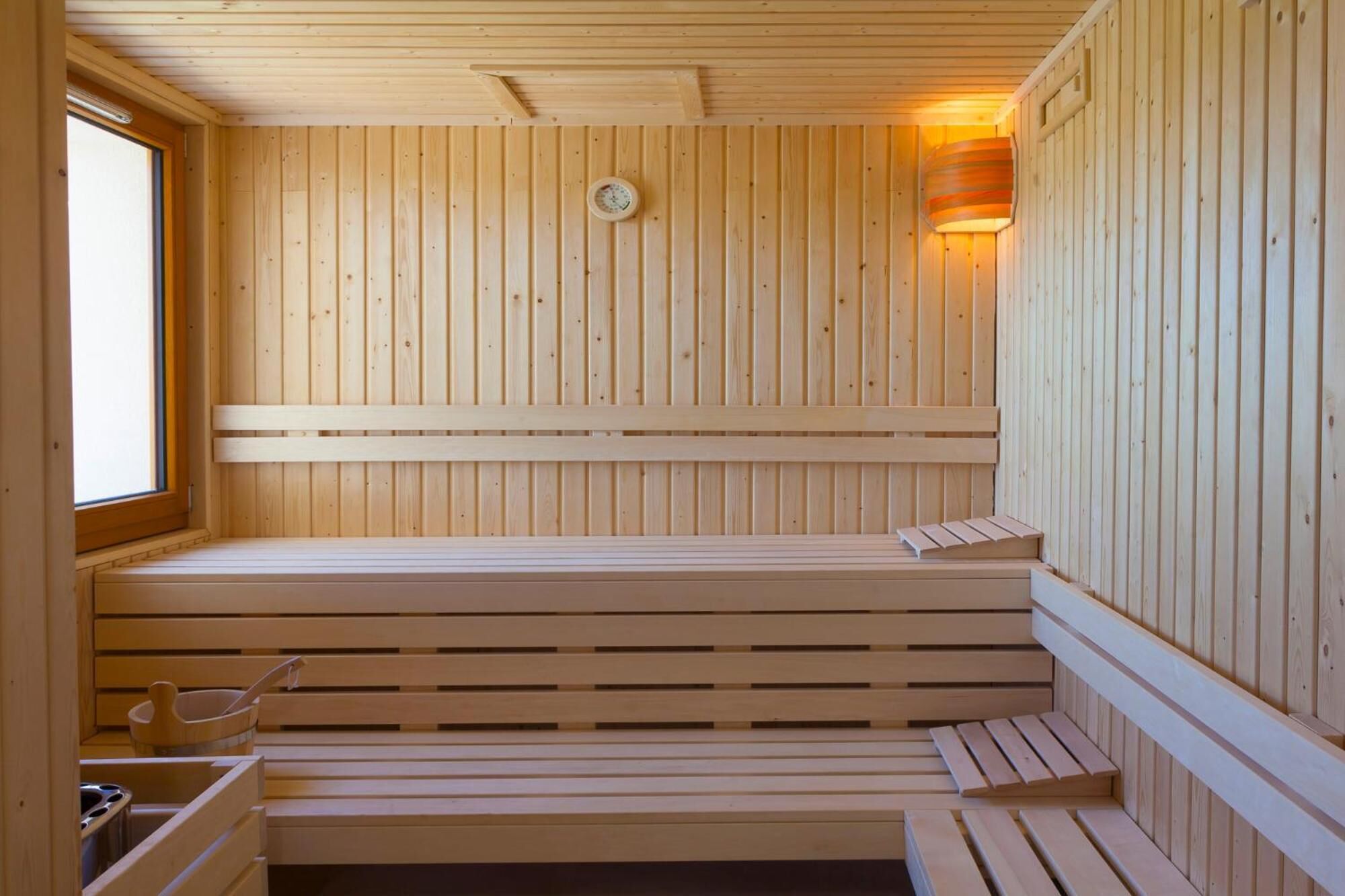 A wooden sauna with benches and a window, featuring a wall-mounted heater and a wall-mounted light fixture.