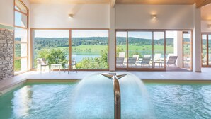 Une piscine intérieure moderne agrémentée d’une cascade, de grandes fenêtres offrant une vue sur un paysage verdoyant et un mur de pierre sur l’un des côtés.