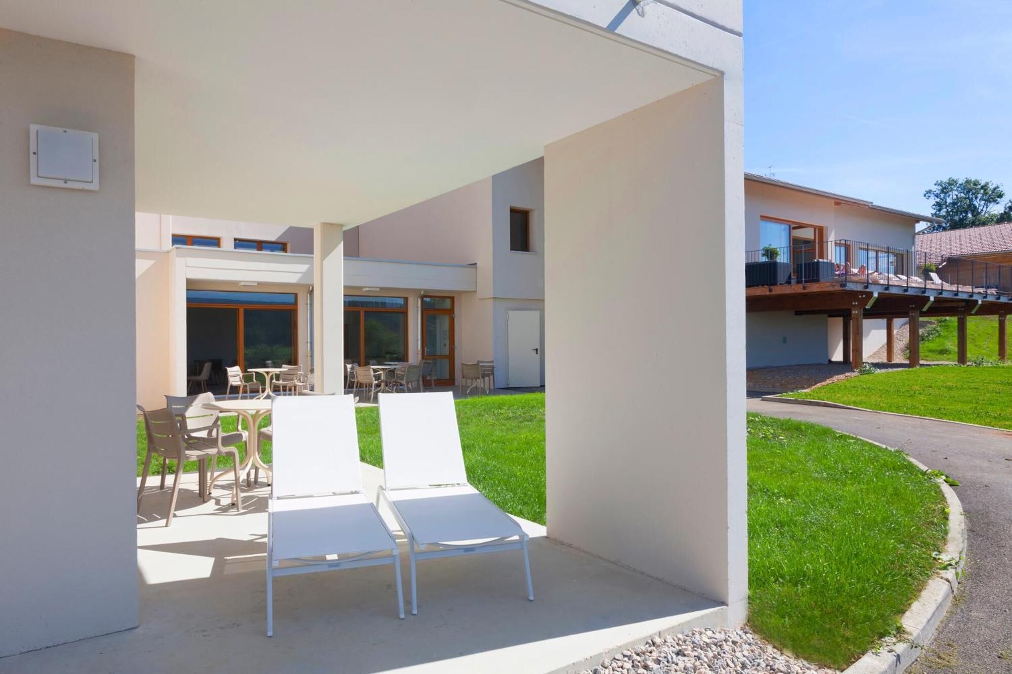 Un espace patio extérieur moderne, avec des chaises longues et des tables blanches, un coin salon abrité et une vue sur un immeuble résidentiel doté d’un balcon.