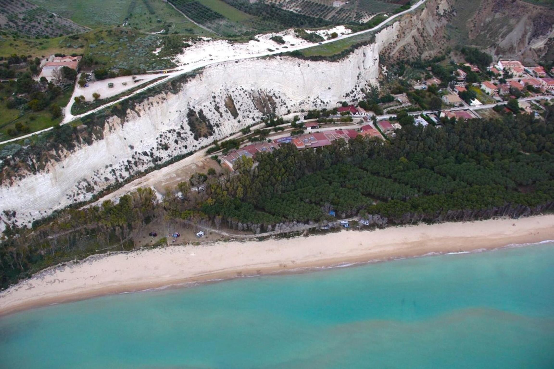 An aerial view of a coastal area with a beach, a cliff, and a small settlement with buildings.