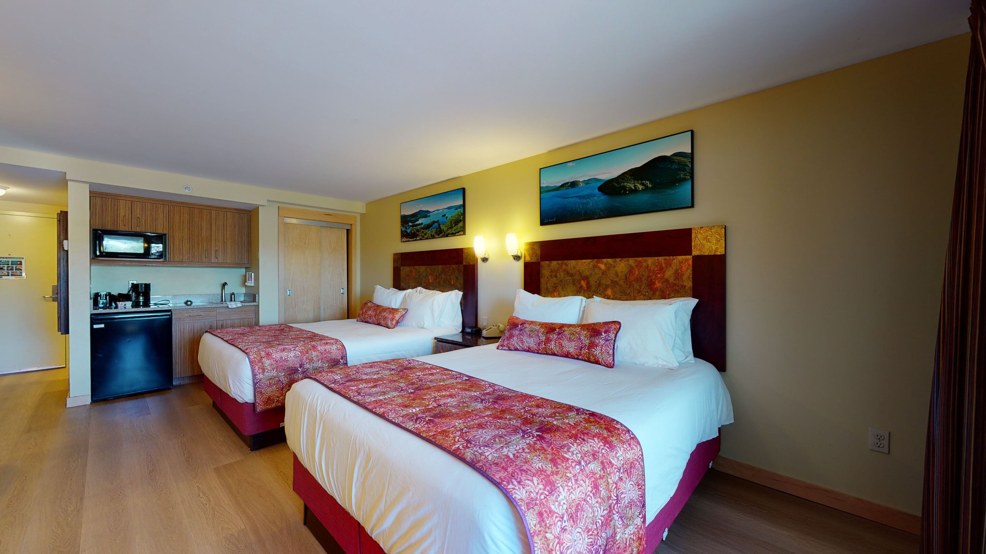 A hotel room with two beds, a kitchenette, and a painting of a landscape.