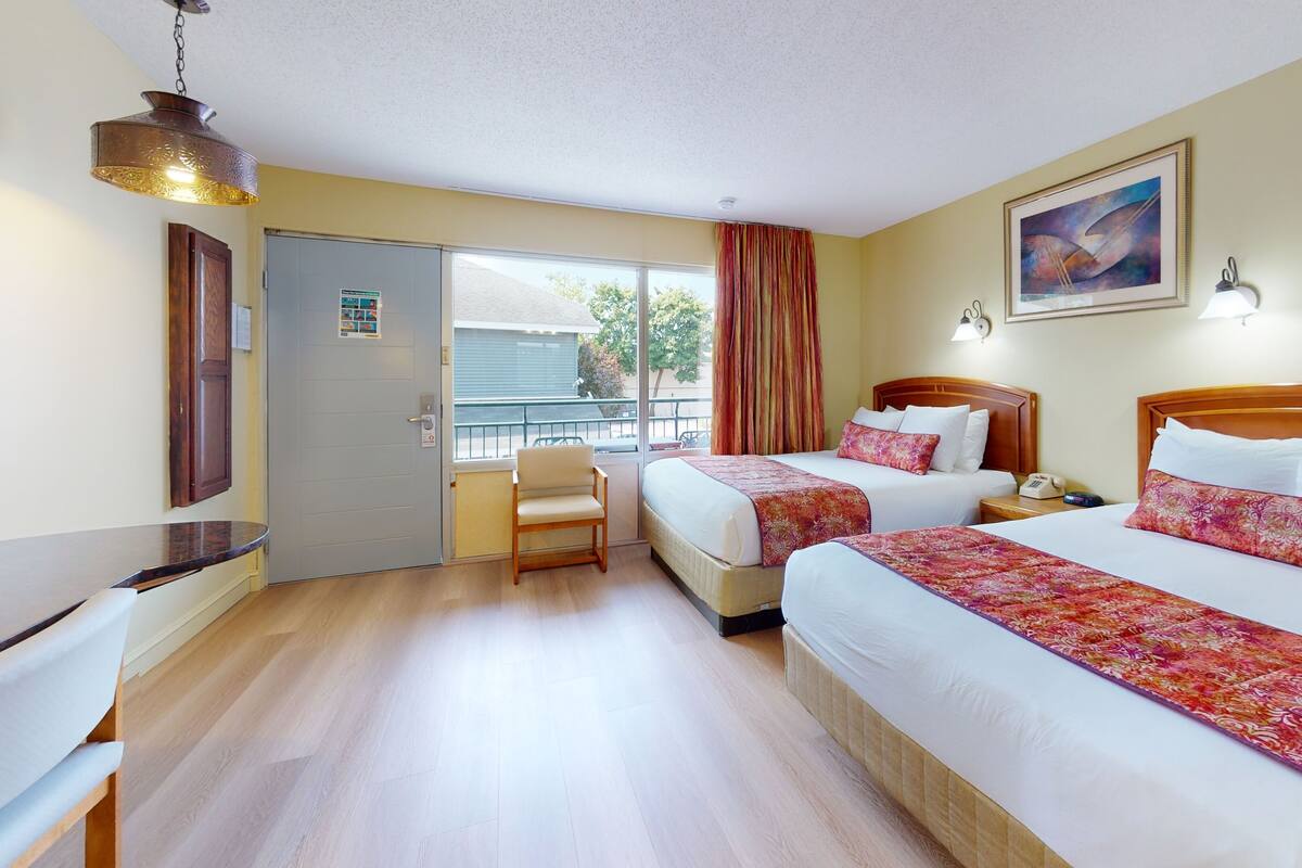 A hotel room with two beds, a wooden headboard, a desk, a chair, a window with curtains, and a painting on the wall.
