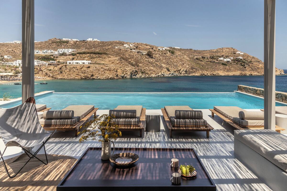 A pool area with lounge chairs and a view of the sea and hillside buildings.