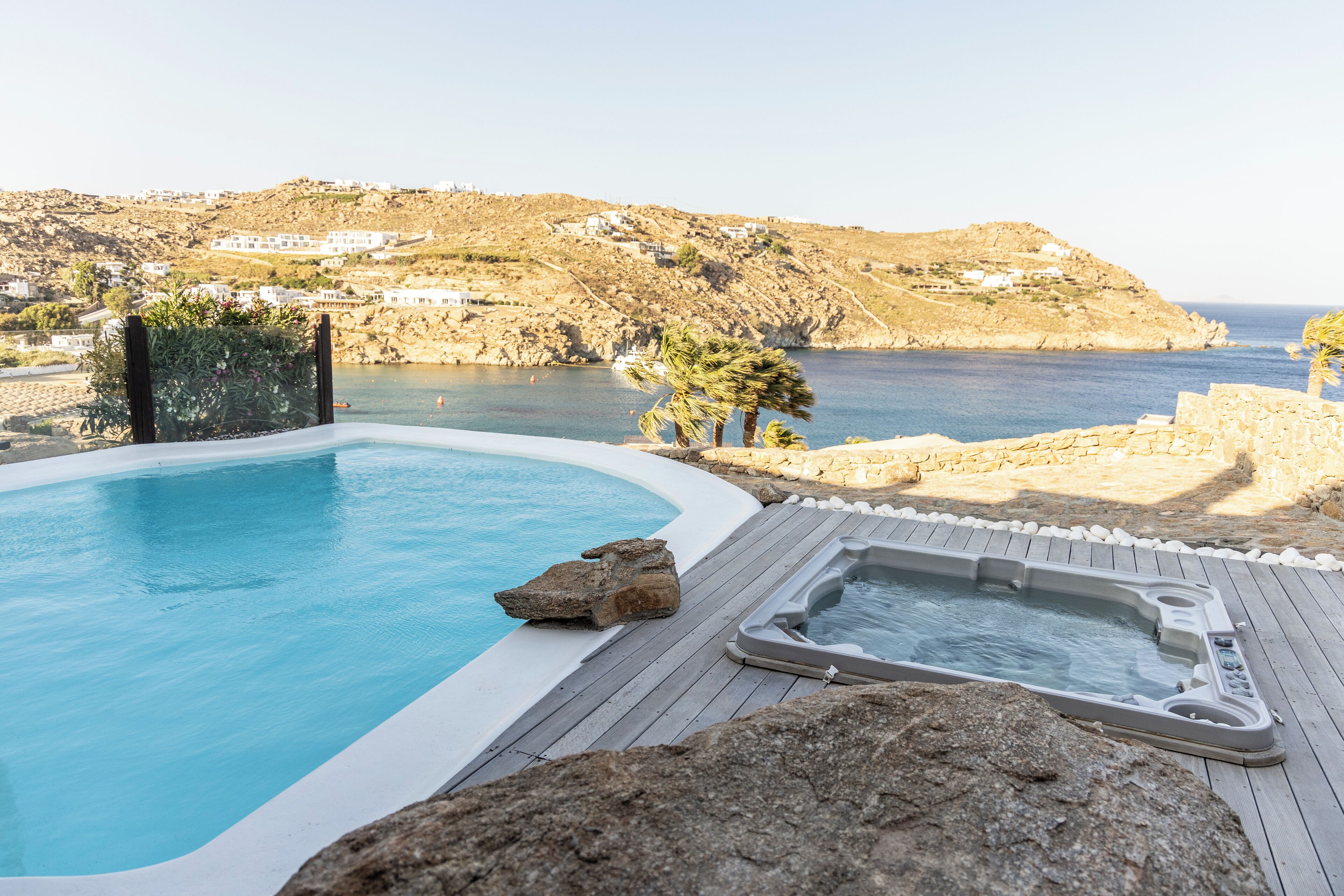 A pool with a hot tub, surrounded by rocks and a wooden deck, overlooking a coastal area with buildings on a hillside.