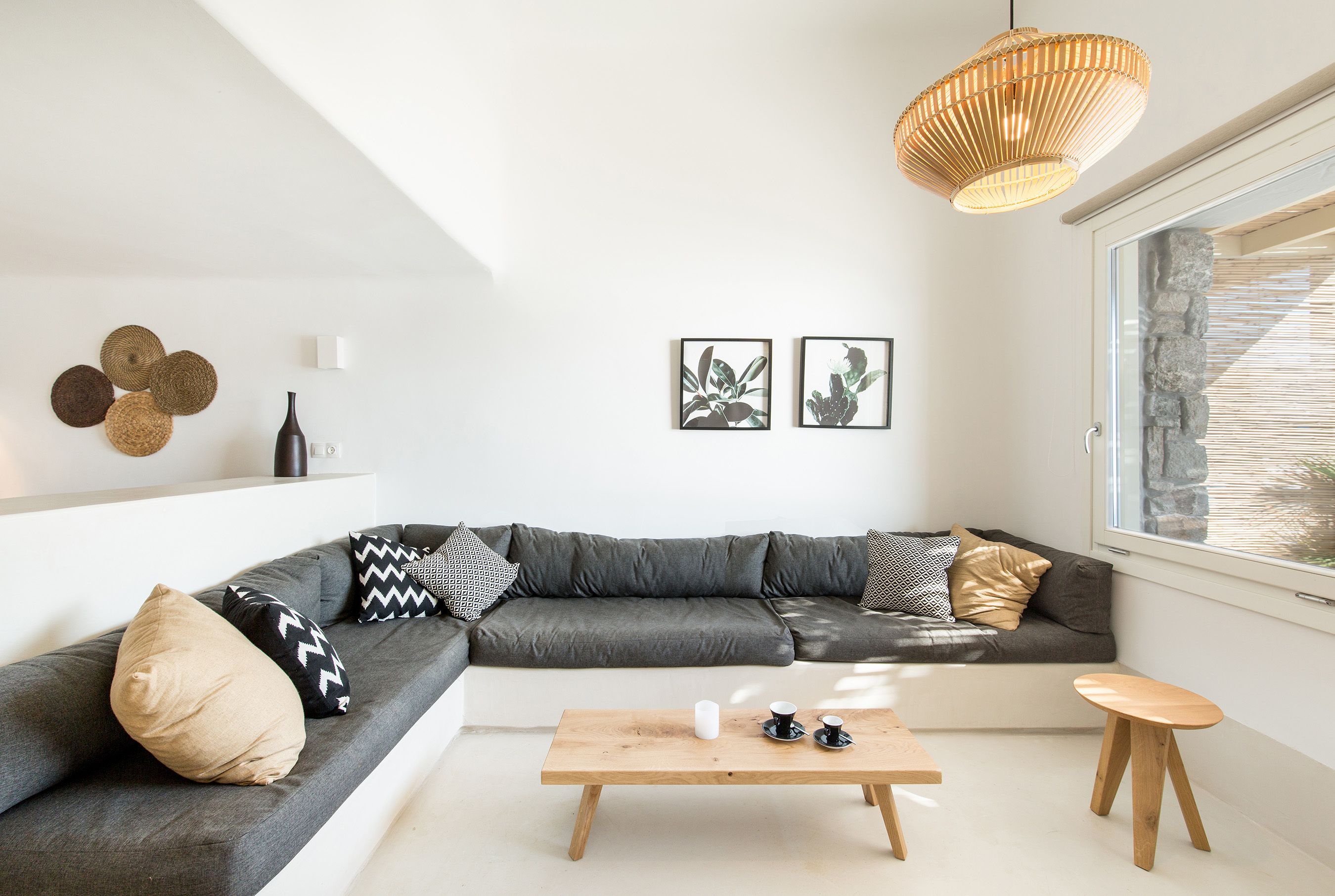 Un salon moderne avec un canapé d’angle gris, une table basse en bois et un petit tabouret en bois. Deux reproductions botaniques encadrées ornent le mur, ainsi qu’un luminaire en fibres naturelles et une fenêtre avec un cadre en pierre.