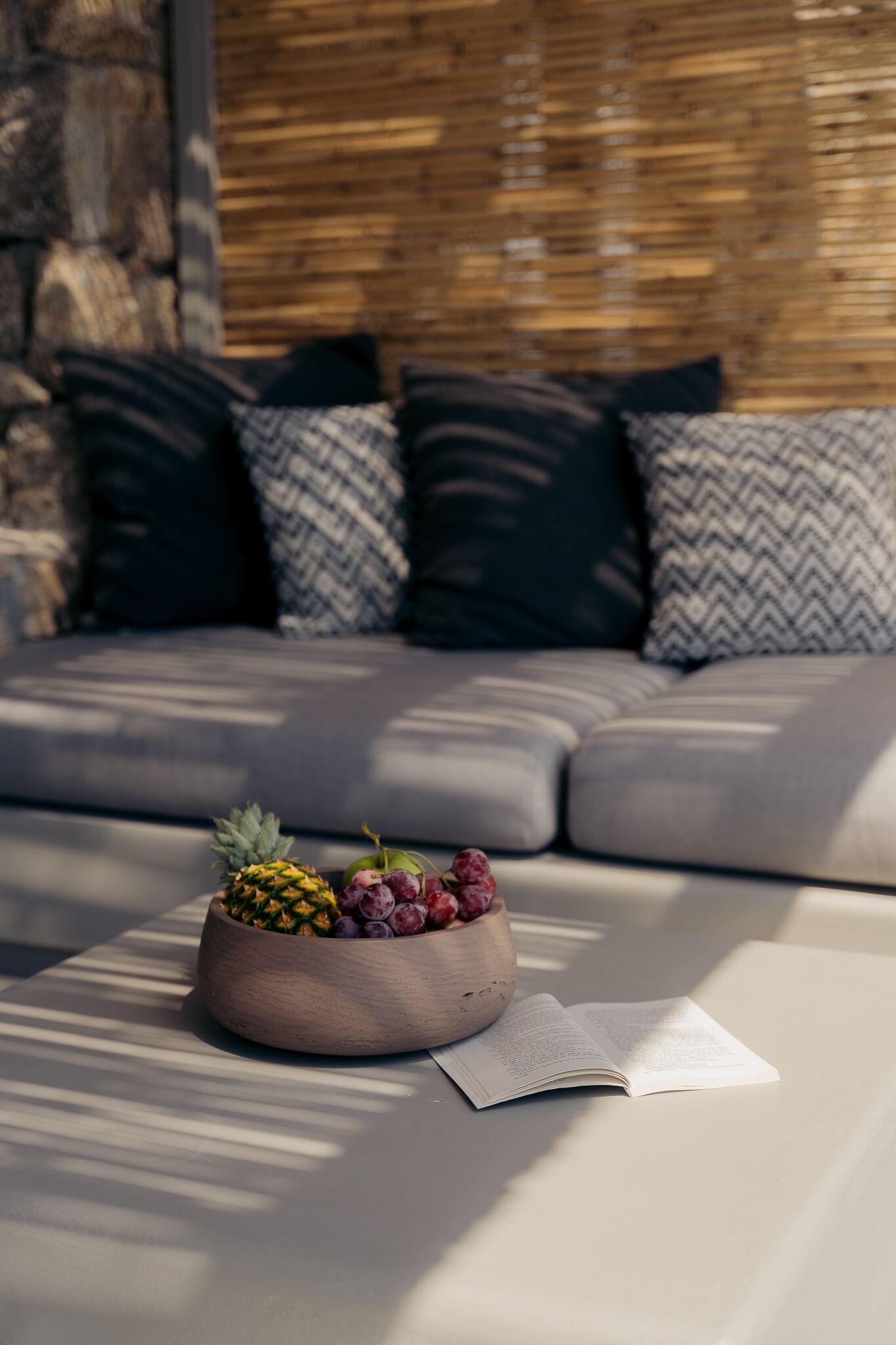 A modern living room with a sofa, cushions, a wooden bowl with fruits, and a book on a table.