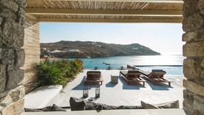 A stone-walled outdoor seating area with lounge chairs and a view of a coastal landscape. - Nimbus My Aktis (Mykonos)