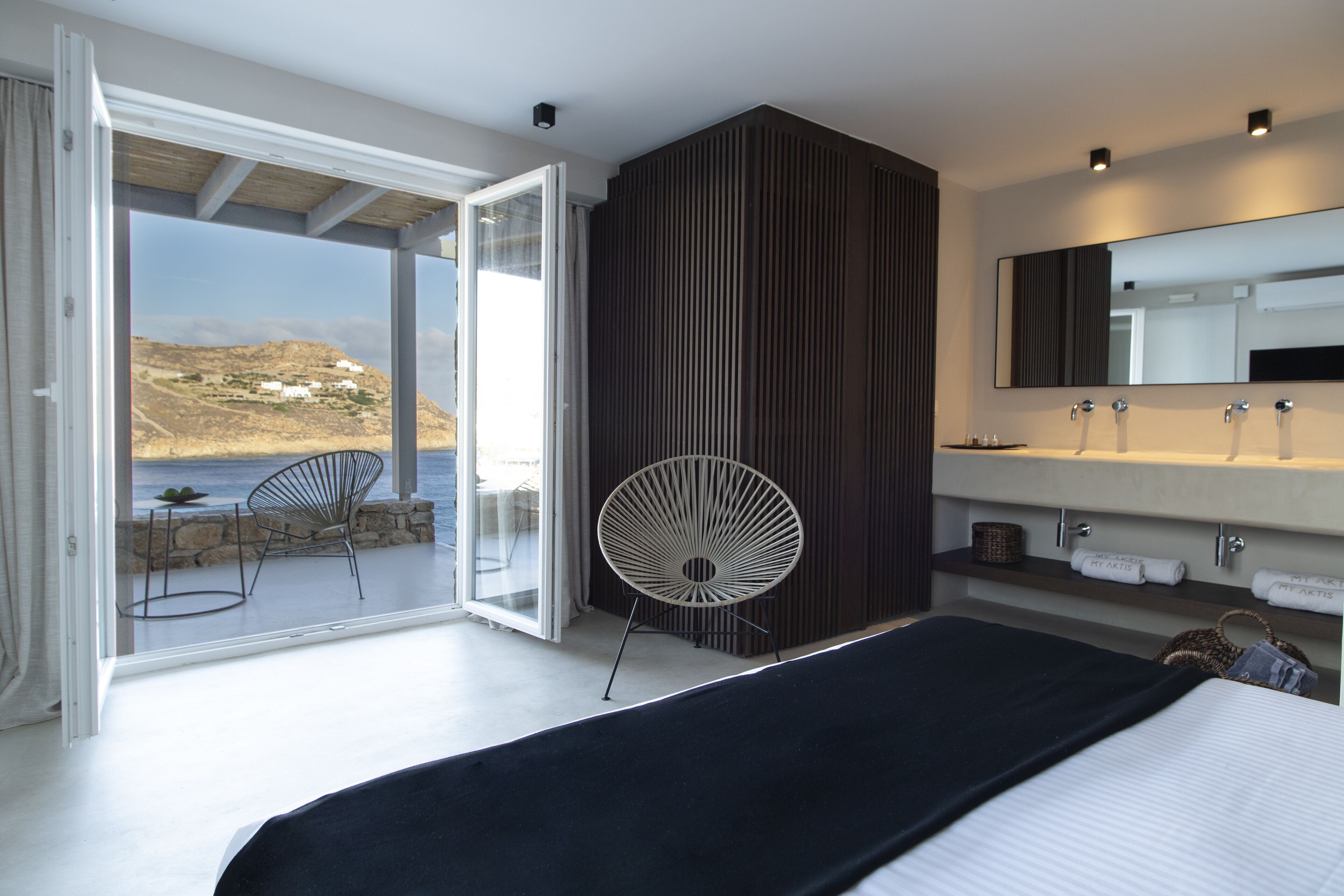 A modern bedroom with a large bed, a bathroom with a mirror and two sinks, a fan, and a view of the sea through an open door.