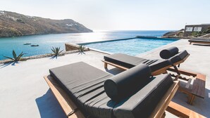 An infinity pool with lounge chairs and a view of the sea. - Nimbus My Aktis (Mykonos)