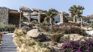 A stone building with a wooden entrance, surrounded by rocks, palm trees, and various plants. - Nimbus My Aktis (Mykonos)
