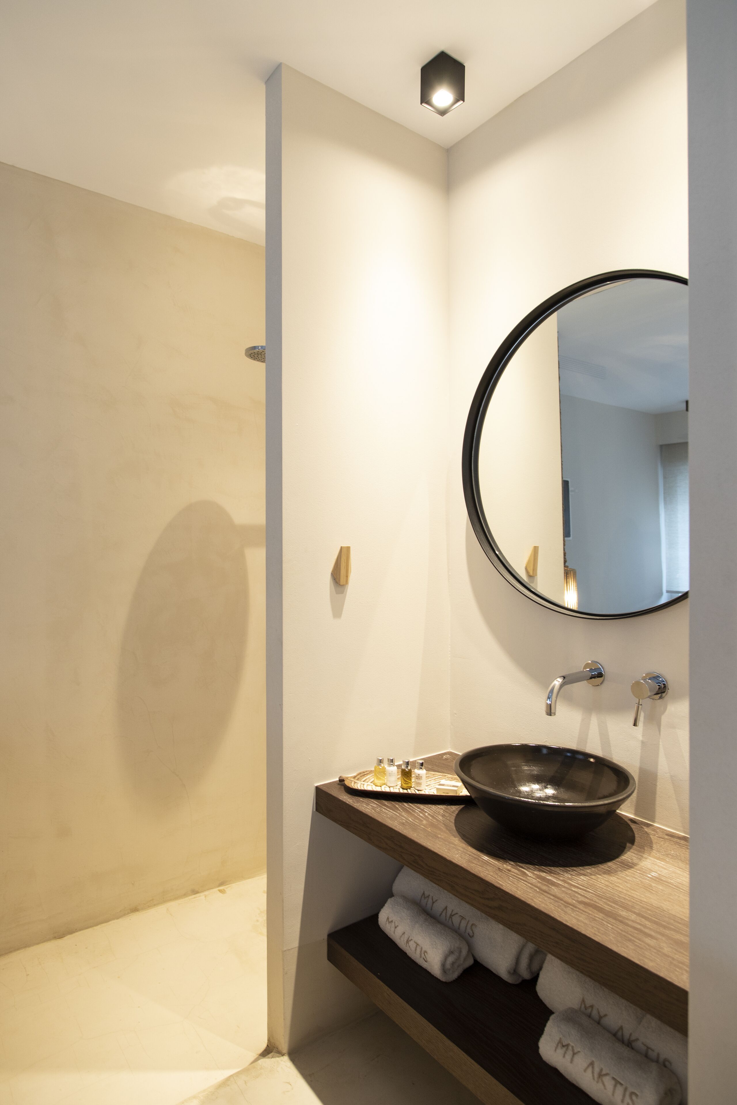 Un bagno moderno con uno specchio rotondo, un lavandino nero e asciugamani ripiegati con cura con la scritta "MY AKTIS" sullo scaffale.