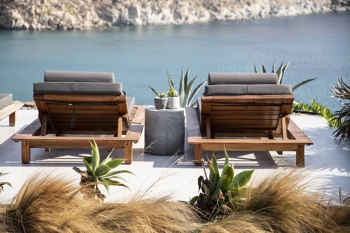 A serene outdoor seating area with two wooden lounge chairs and a round table, surrounded by potted plants and overlooking a calm body of water.