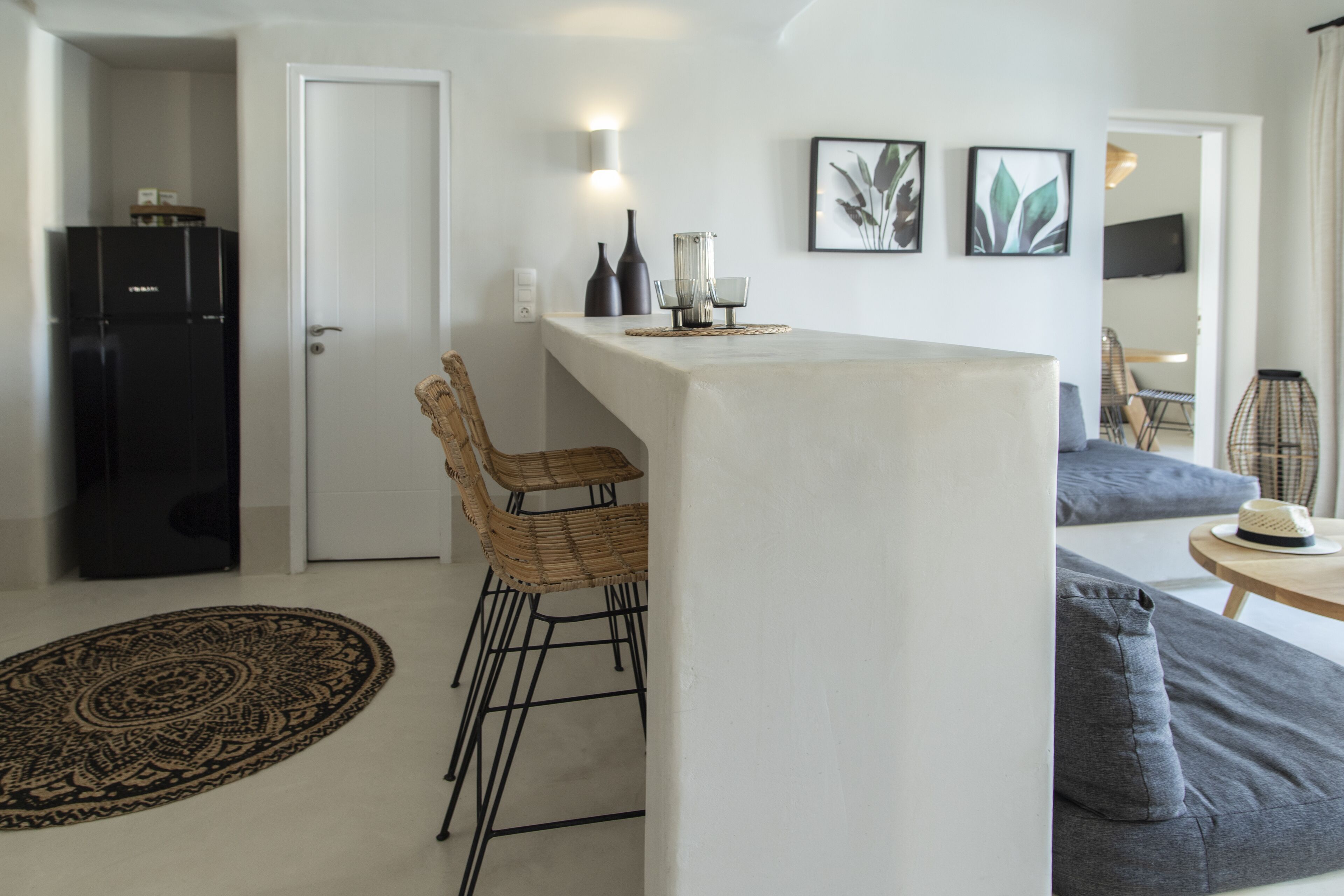 A modern kitchen with a white countertop, black refrigerator, and wicker chairs. There are framed pictures on the wall and a round rug on the floor.
