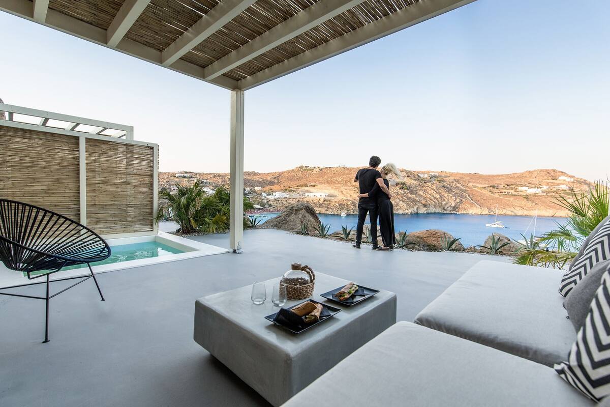 A modern outdoor seating area with a pool, a dining table with plates, and a view of a coastal landscape.