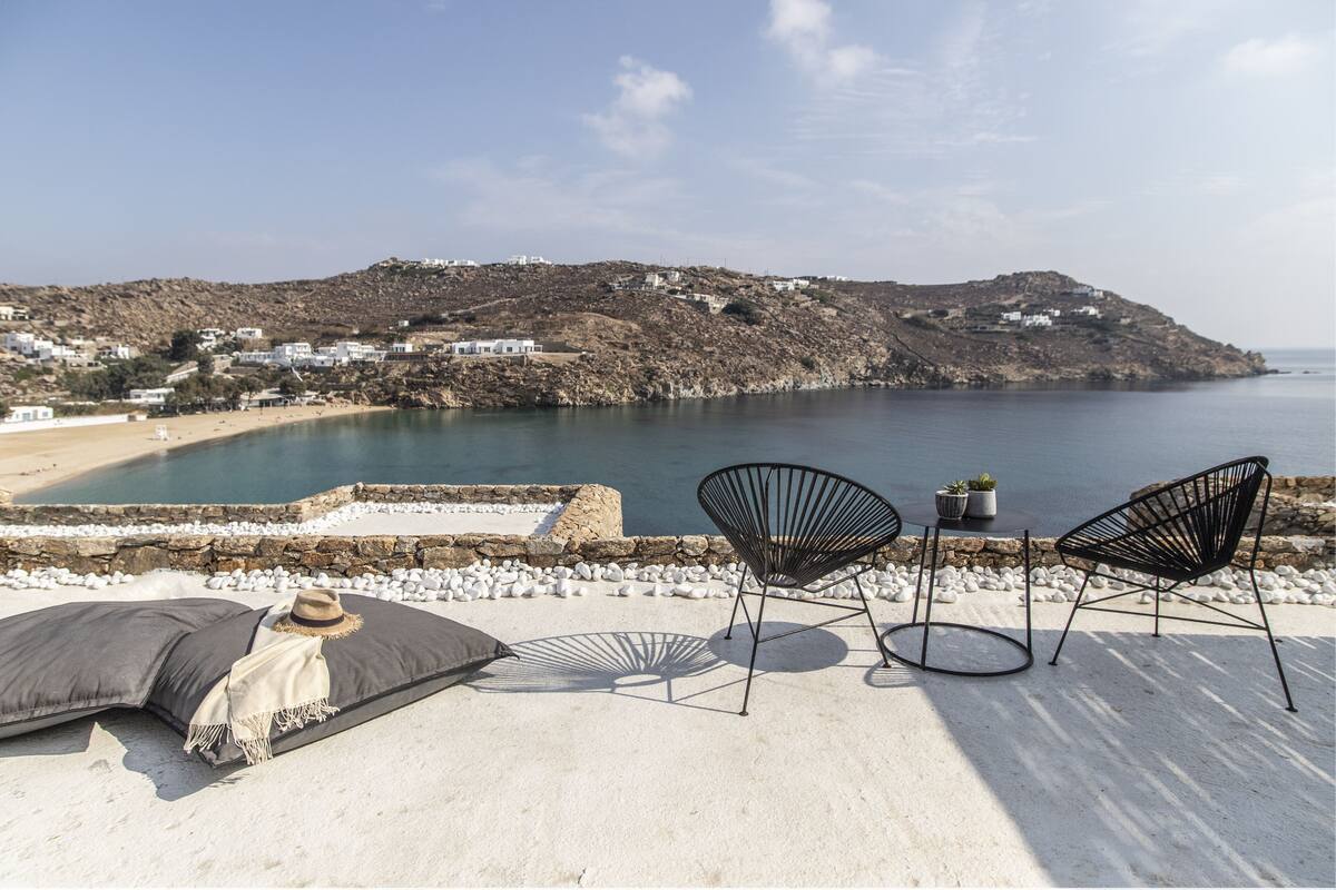 A beachside terrace with a view of the sea and a hillside. There are two black chairs and a table with a plant.
