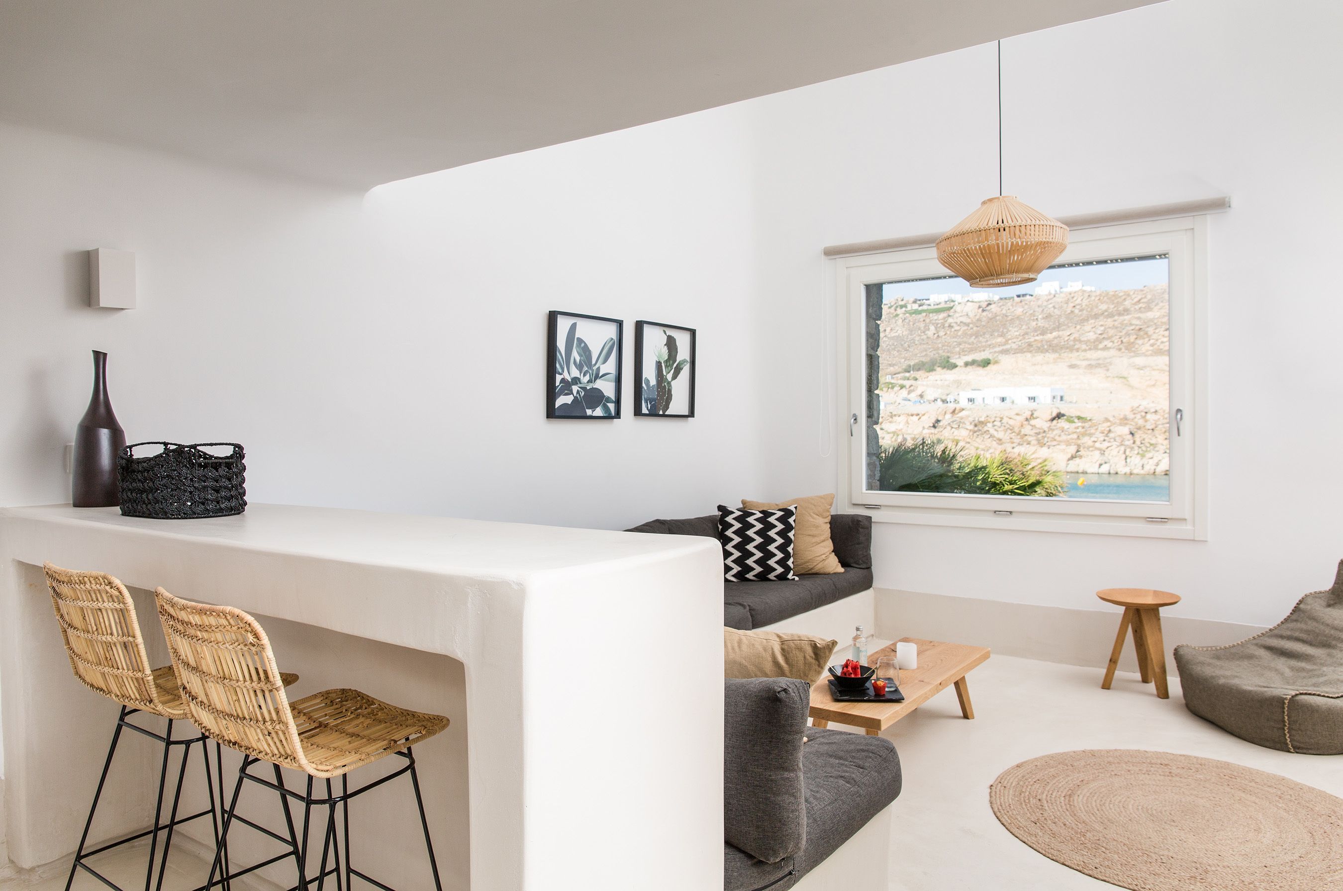 A modern living room with a white sofa, a grey couch, and a dining area with wicker chairs. There is a large window with a view of a rocky landscape.