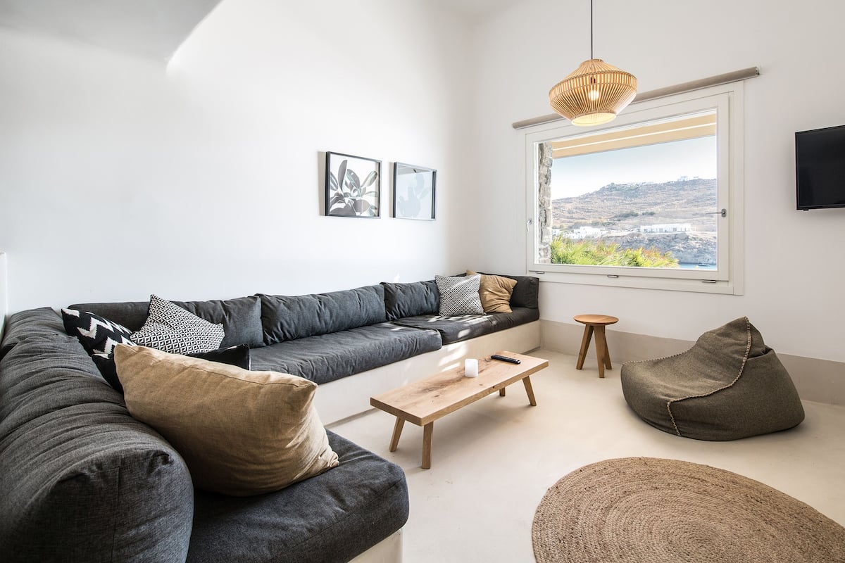 A modern living room with a grey sofa, a wooden coffee table, a bean bag, and a view of a coastal landscape through a large window.
