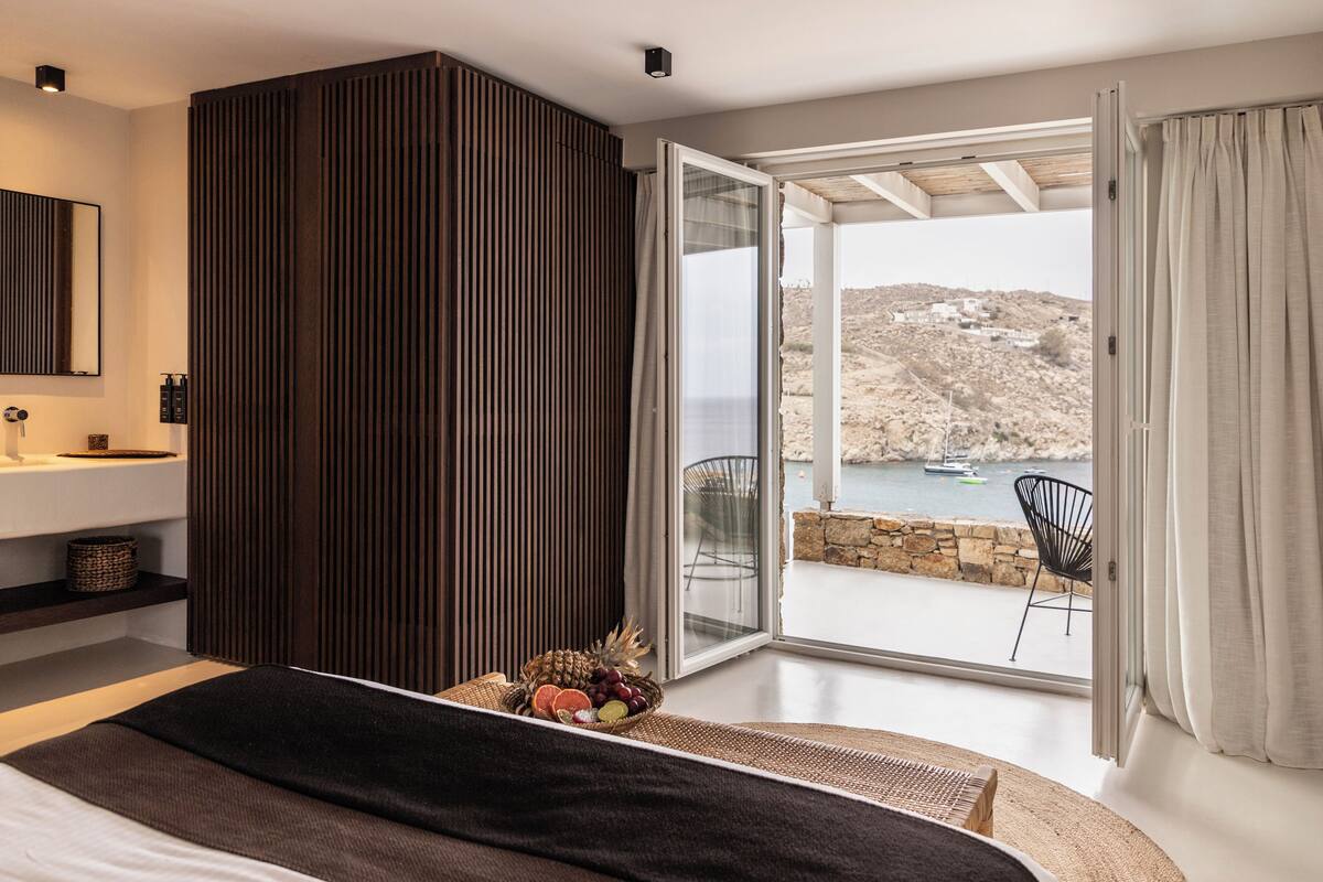 A bedroom with a bed, a fruit basket, a view of a rocky landscape through a sliding glass door, and a stone wall.
