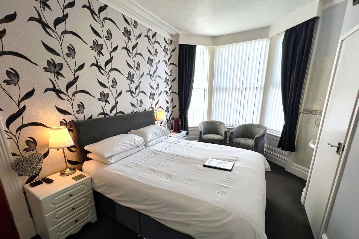 A hotel room with a large bed, two armchairs, a nightstand with a lamp, and a window with curtains.