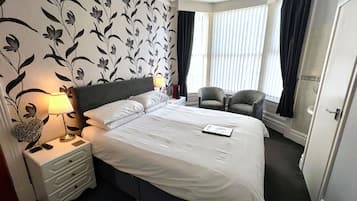 A hotel room with a large bed, two armchairs, a nightstand with a lamp, and a window with curtains.