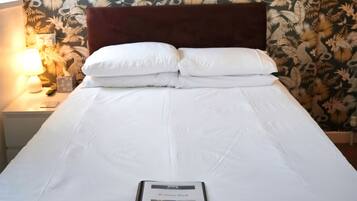 Een net opgemaakt bed met witte lakens en kussens, een bord met welkomstboodschap in een lijst en behang met een bloemenmotief.