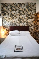A neatly made bed with white linens and pillows, a framed welcome sign, and a floral-patterned wallpaper.