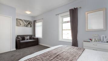 Een slaapkamer met een bed, een nachtkastje, een dressoir, een raam met gordijnen, een aan de muur hangend kunstwerk, en een bank.