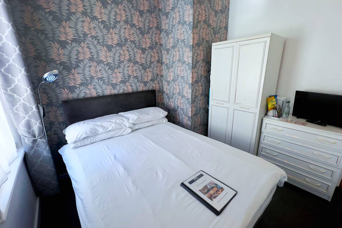 A neatly made bed with white linens, a bedside lamp, a wardrobe, a television, and a magazine on the bed.