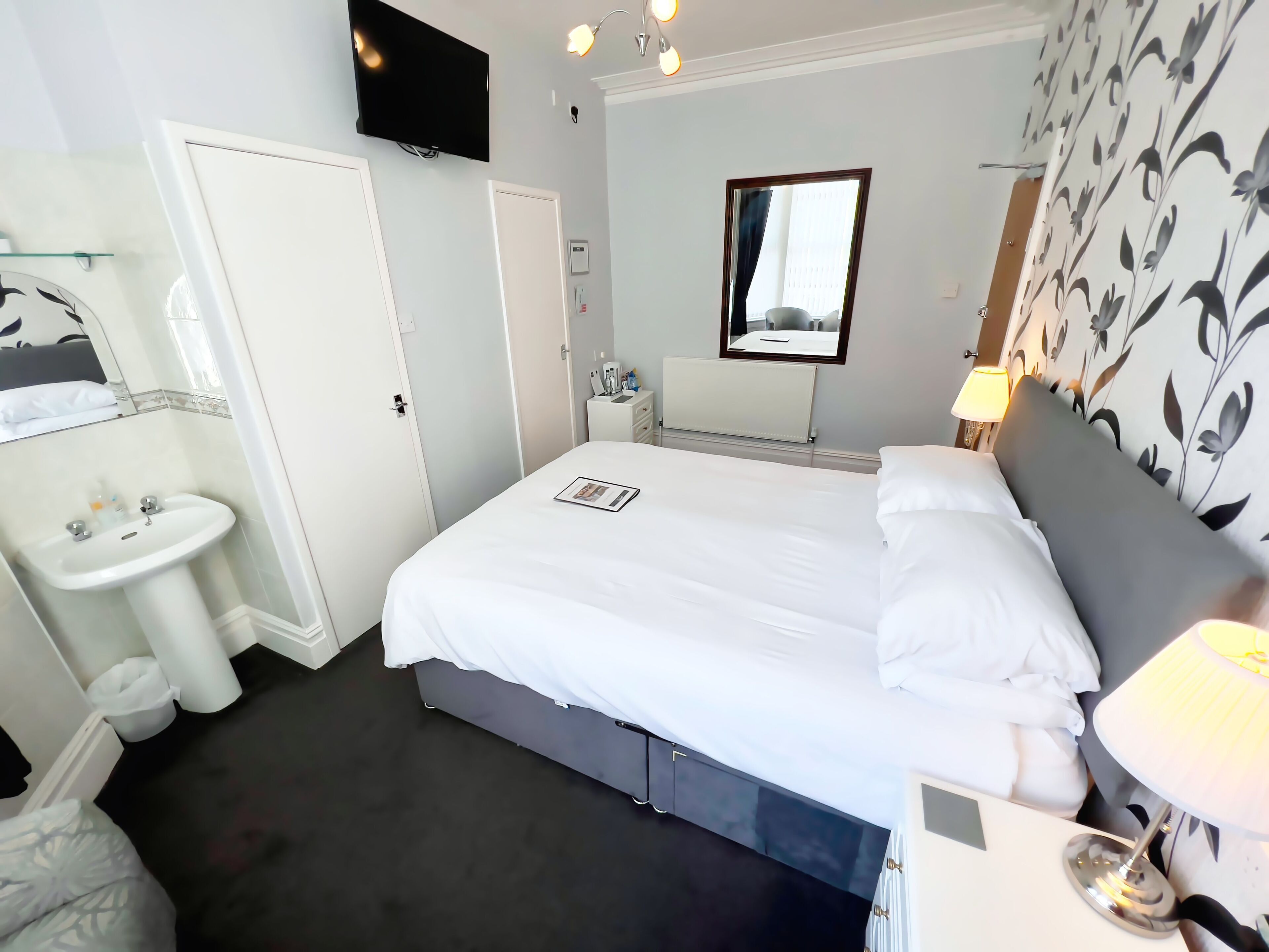 Une chambre d’hôtel comprenant un lit, une table de chevet avec une lampe, un téléviseur fixé au mur, une salle de bain avec un lavabo et un miroir.