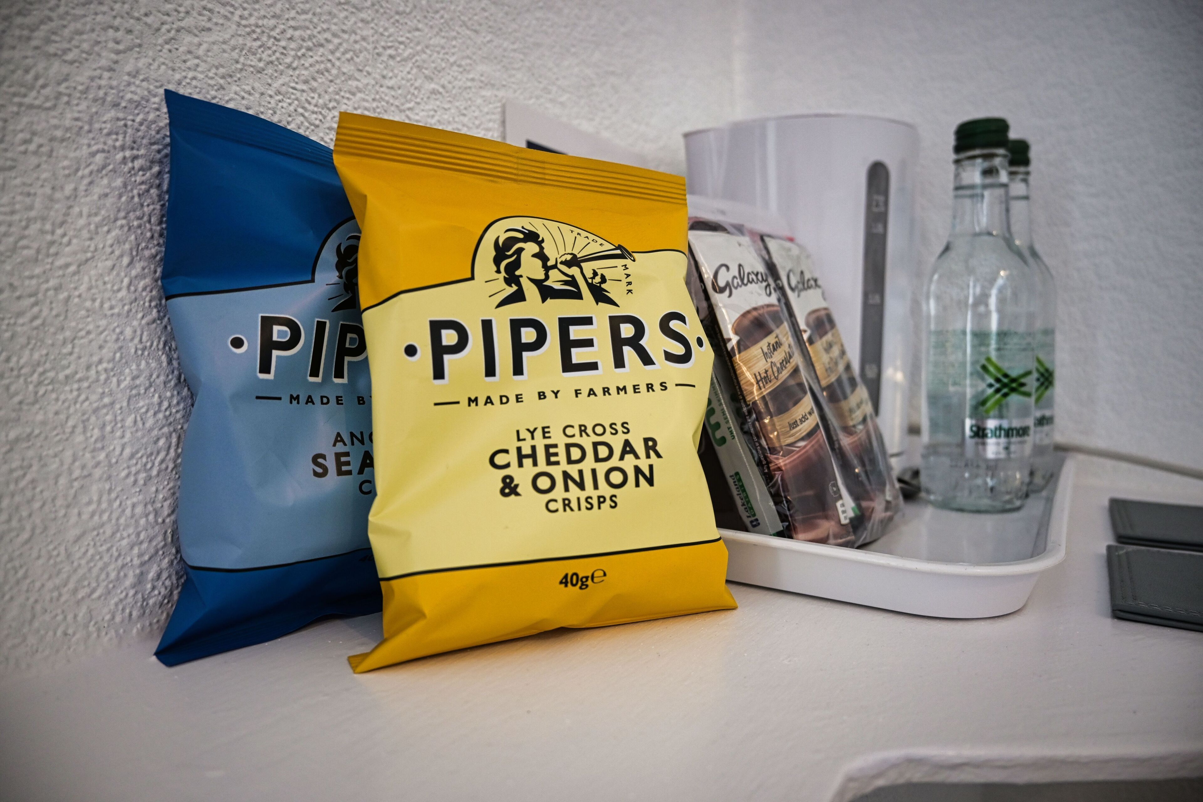 A kitchen counter with a bag of Pipers Lye Cross Cheddar & Onion Crisps, a coffee maker, and two bottles of water.