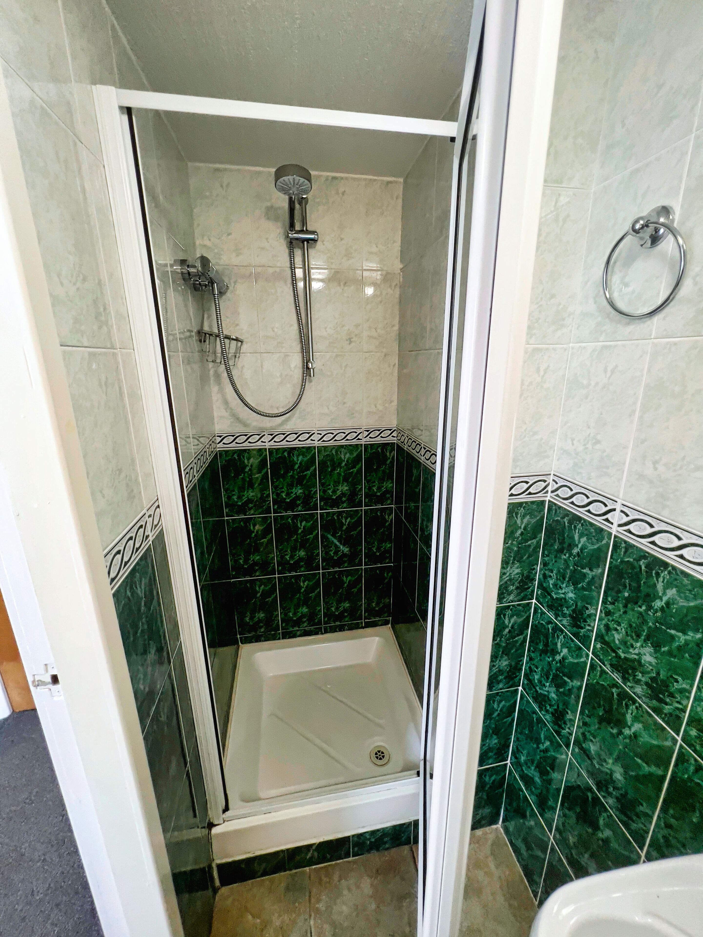 Una ducha a nivel con puerta de vidrio, un cabezal de ducha manual y un grifo empotrado en la pared. El área de la ducha tiene azulejos de mármol verde con detalles blancos.