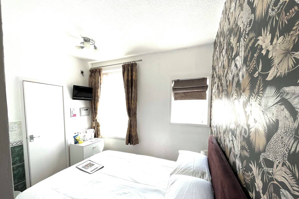 A hotel room with a bed, a desk with a chair, a window with curtains, a wall-mounted TV, and a small window with a view of the outside.