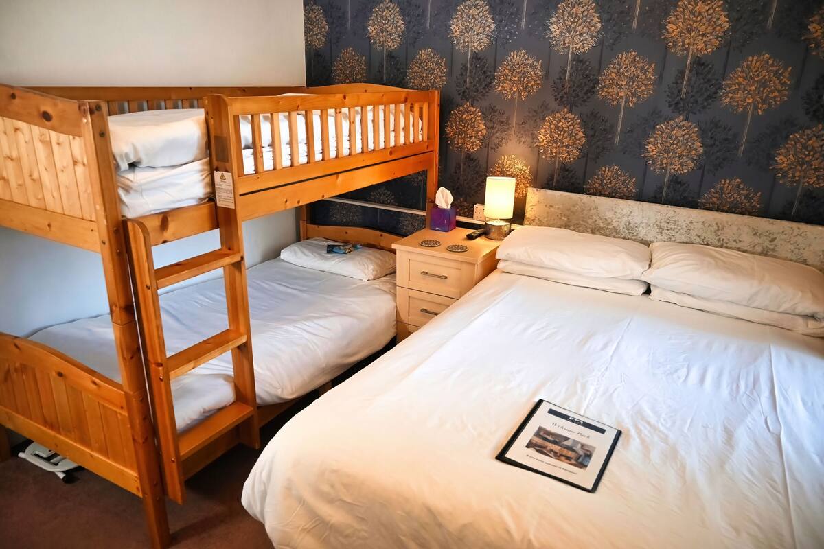 A room with a bunk bed, a single bed, a nightstand with a lamp, and a wall with a floral pattern.