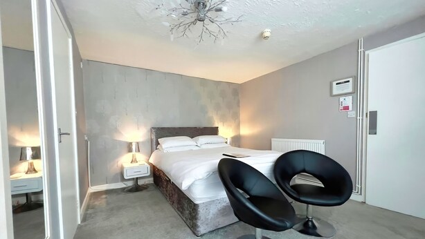 A bedroom with a bed, bedside lamps, a radiator, and two black chairs. - Bella Vista Lodge (Blackpool)