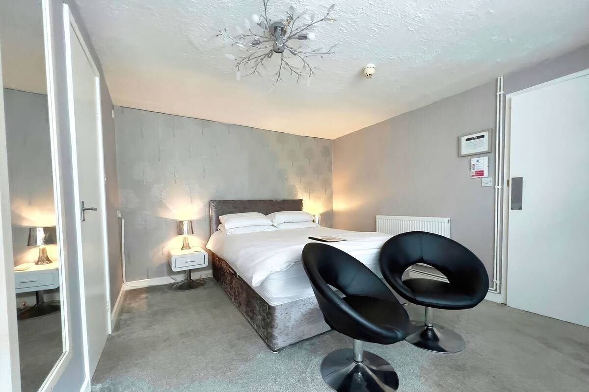 A bedroom with a bed, bedside lamps, a radiator, and two black chairs.