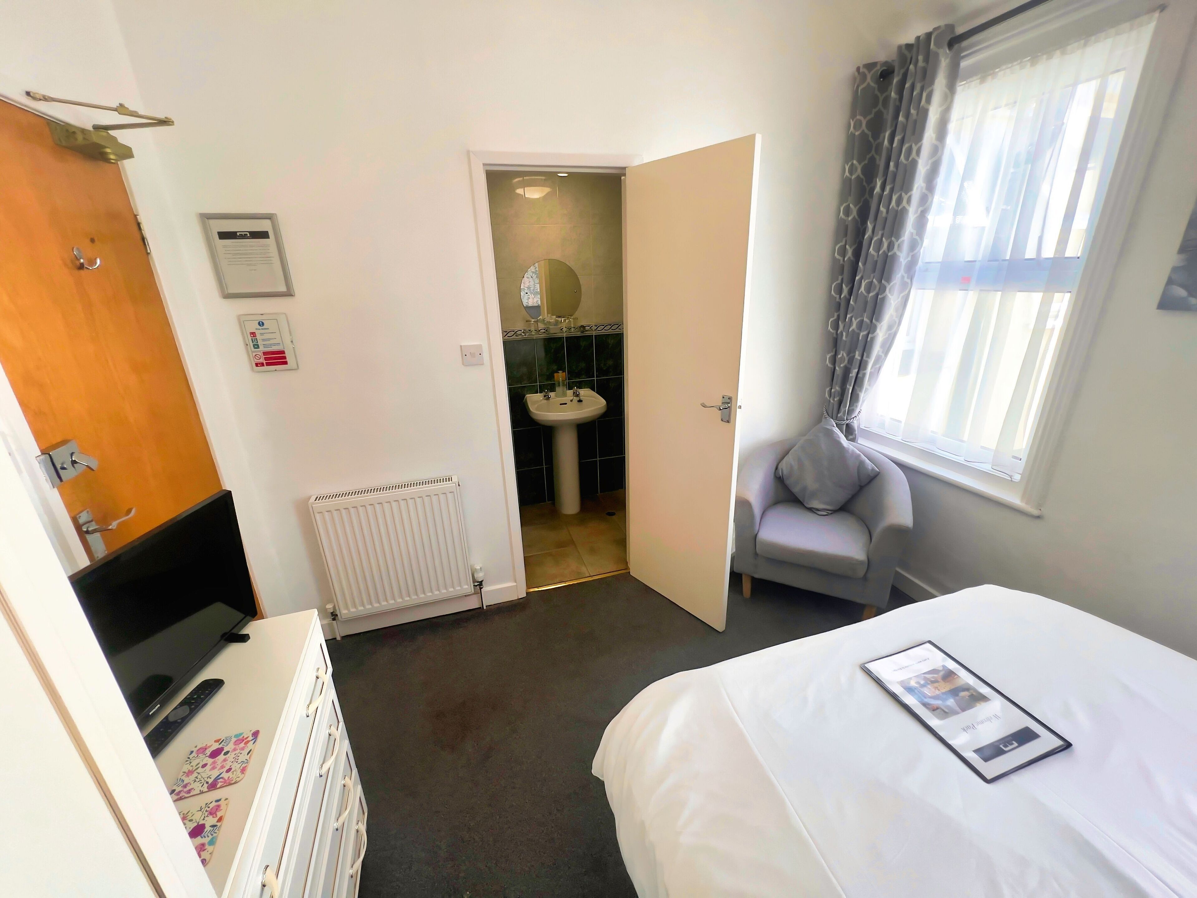 Una camera d'albergo con un letto, una sedia, un termosifone, una porta e una finestra con tende.